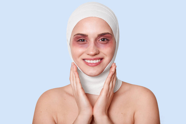 indoor shot of cheerful patient being satisfied after cosmetic procedures for appearance improvement , has bruises under eyes, touches cheeks with charming smile, stands against blue studio wall. - Foto, imagen