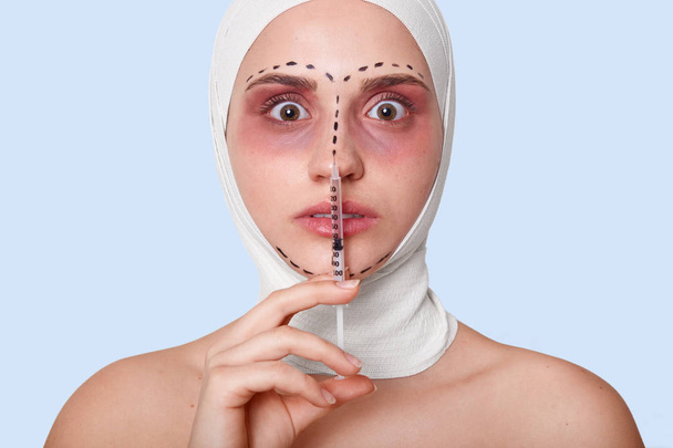 Shot of shocked scrared woman staring with bugged eyes, ready for recieves injection, afraids of plastic surgery, has marked lines on problematic zones of face, isolated over blue studio background - Photo, Image