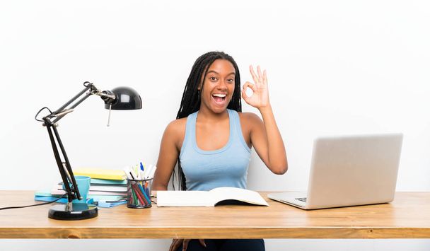 Αφρικανική αμερικανική έφηβος μαθήτρια με μακρύ πλεκτό μαλλιά στο χώρο εργασίας της έκπληκτος και δείχνοντας OK πινακίδα - Φωτογραφία, εικόνα