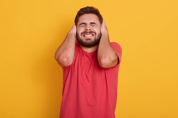 Image of handsome bearded male wearing redcasual t shirt, attractive man posing with closed eyes and ears, hear loud noise, guy standing isolated over yellow studio background. People concept. - Fotografie, Obrázek