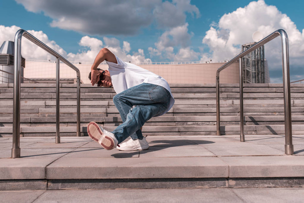 a young guy a dancer, makes a lower break, dances in motion in the summer in the city against the background of steps and a building, jeans, sunglasses, a white T-shirt - Foto, Bild