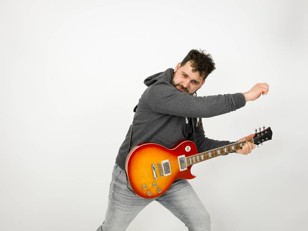 cool man with black hair and beard wearing grey hoodie playing electric guitar in front of white background - Fotoğraf, Görsel