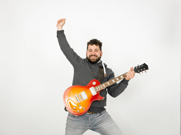 cool man with black hair and beard wearing grey hoodie playing electric guitar in front of white background - Фото, изображение