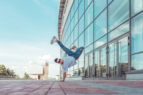 strong sporty man dancing break dance, standing one arm, young guy, free space for text motivation lifestyle, summer city against background glass building window, jeans, sunglasses, a white T-shirt - Фото, зображення