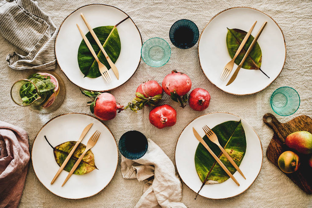 Fall or Autumn table styling for holiday dinner. Flat-lay of dinnerware with fruit and fallen leaves for decoration over beige linen tablecloth, top view. Preparation for Thanksgiving day or Christmas - Foto, Bild