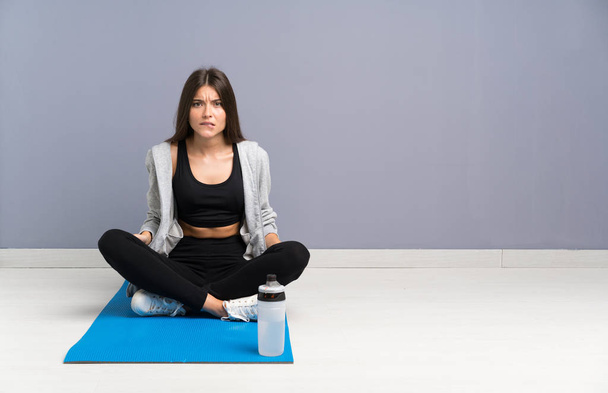 Young sport woman sitting on the floor with mat having doubts and with confuse face expression - Foto, afbeelding