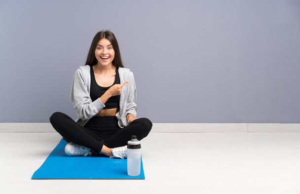 Young sport woman sitting on the floor with mat pointing finger to the side - Photo, Image