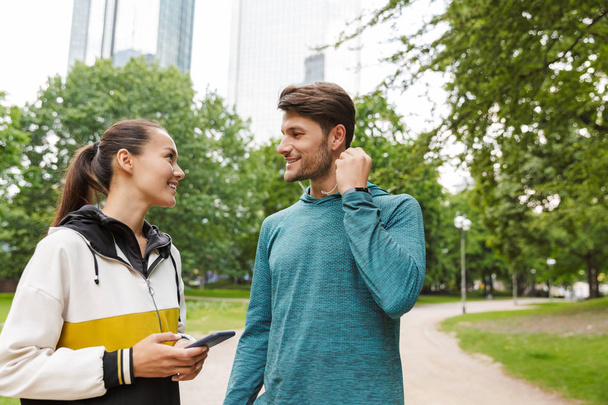 Photo of athletic smiling man and woman using cellphone and earphones while talking in city park - Photo, image
