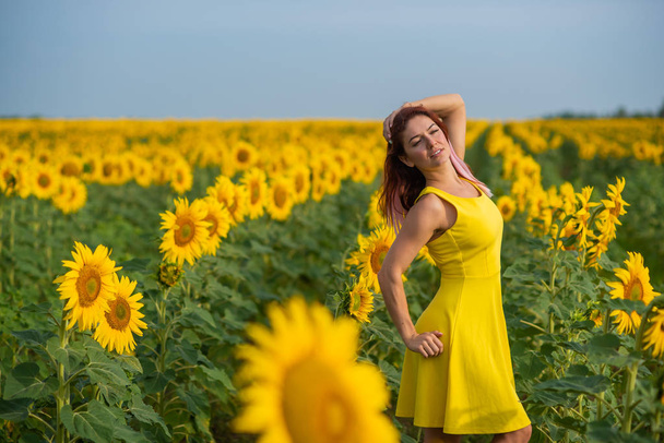 A red-haired woman in a yellow dress is standing in a field of sunflowers. Beautiful girl in a skirt sun enjoys a cloudless day in the countryside. Pink locks of hair. - Photo, Image