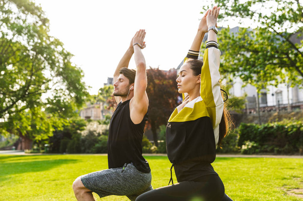 Photo of young focused man and woman doing yoga exercises with eyes closed while working out in city park - Photo, Image