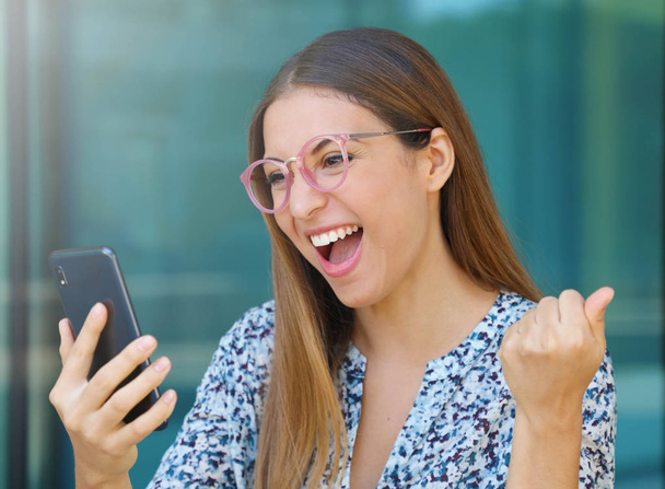Vzrušená mladá žena s brýlemi kontroluje svůj elegantní telefon venku - Fotografie, Obrázek