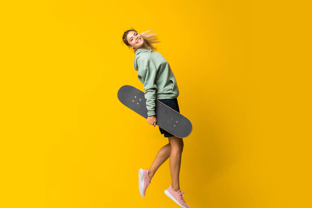 Blonde teenager skater girl jumping over isolated yellow background - Фото, изображение