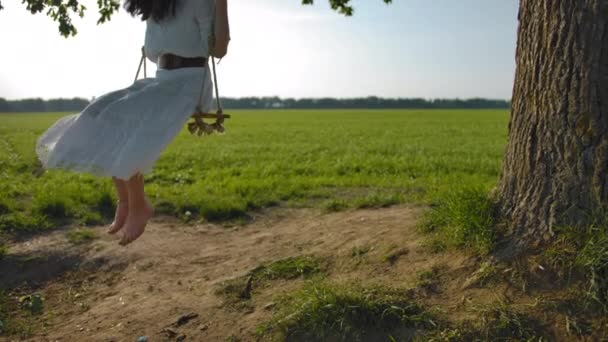 Jonge Barefoot meisje in een mooie witte rustieke jurk is swingend op een schommel in het open veld. - Video
