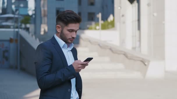 Νέος όμορφος επιχειρηματίας στέκεται κοντά στο μεγάλο κτίριο γραφείων. Δακτυλογράφηση ενός μηνύματος στο smartphone του. Επιχειρηματικός τρόπος ζωής. - Πλάνα, βίντεο
