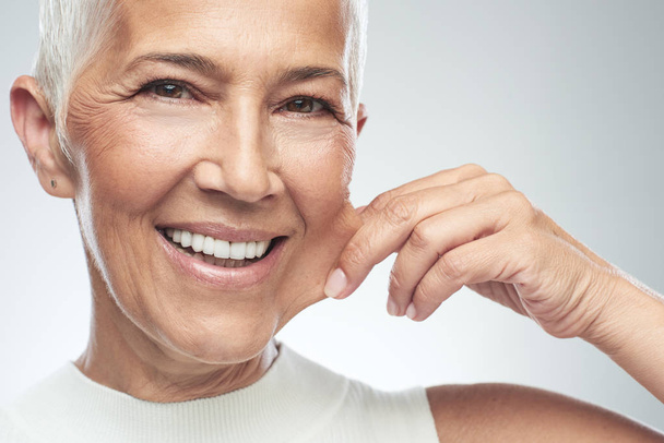 Gorgeous smiling Caucasian senior woman with short gray hair pinching her cheek. Beauty photography. - Photo, Image