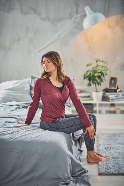 charmante kaukasische Brünette sitzt auf dem Bett im Schlafzimmer und macht Yoga-Übungen. - Foto, Bild