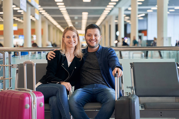 Image of happy woman and man in love looking at camera while sitting in waiting room at airport. - Foto, Imagen
