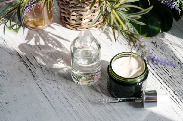 Cosmetic cream container and a bootle of serum with green herbal leaves on white table - Fotografie, Obrázek