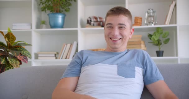 Closeup portrait of adult attractive caucasian man watching TV laughing cheerfully holding remote control sitting on couch indoors - Footage, Video