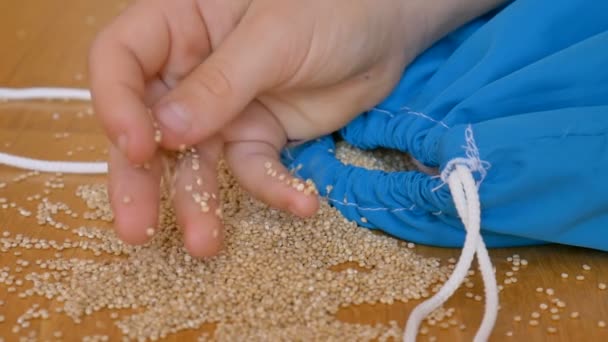Kind handen spelen met witte quinoa zaden in blauwe herbruikbare pakket op houten achtergrond. Eco-verpakking en gezond eetconcept - Video