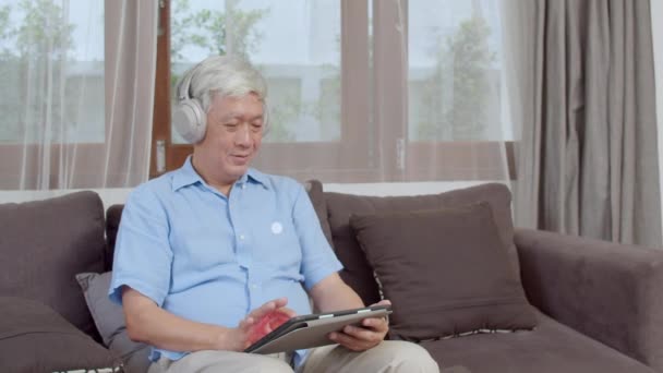 Asian senior man relax at home. Asian older male happy wear headphone using tablet listening podcast while lying on sofa in living room at home concept. - Séquence, vidéo