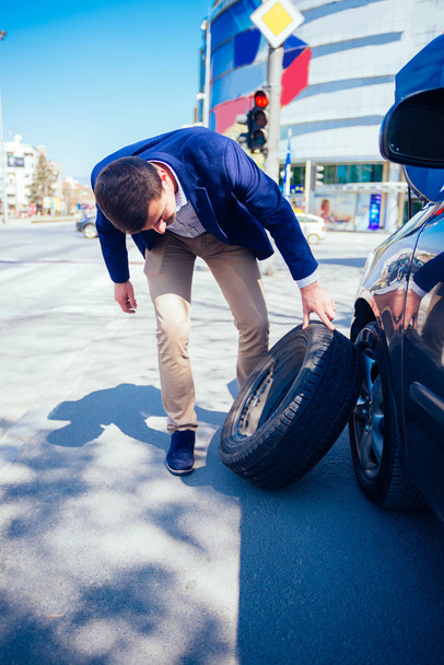 Un homme d'affaires fatigué par une journée ensoleillée sort un pneu de sa voiture afin de pouvoir changer son pneu crevé
. - Photo, image
