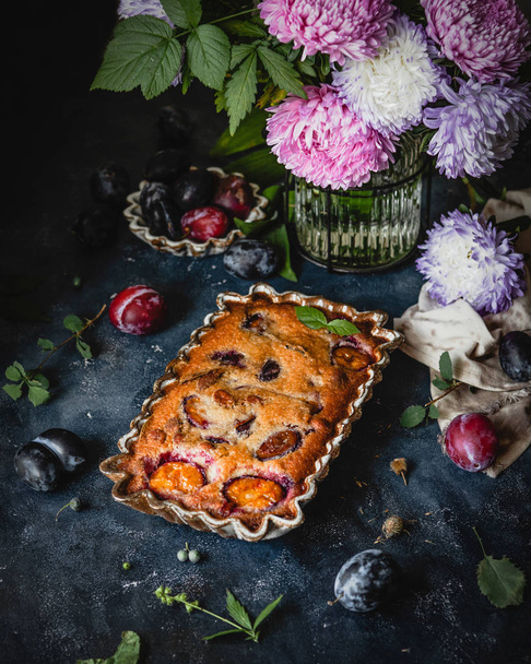 baked plum pie in ceramic mold among fresh plums and flowers on blue background - Photo, Image