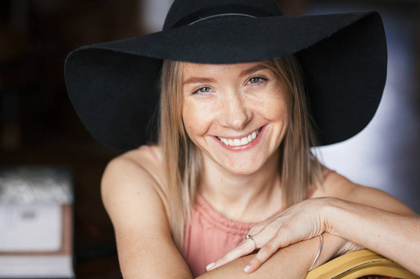 Close warm romantic portrait of a beautiful happy smiling young blonde woman with freckles in ash rose color dress and black felt hat. Girl sitting indoors - Fotoğraf, Görsel