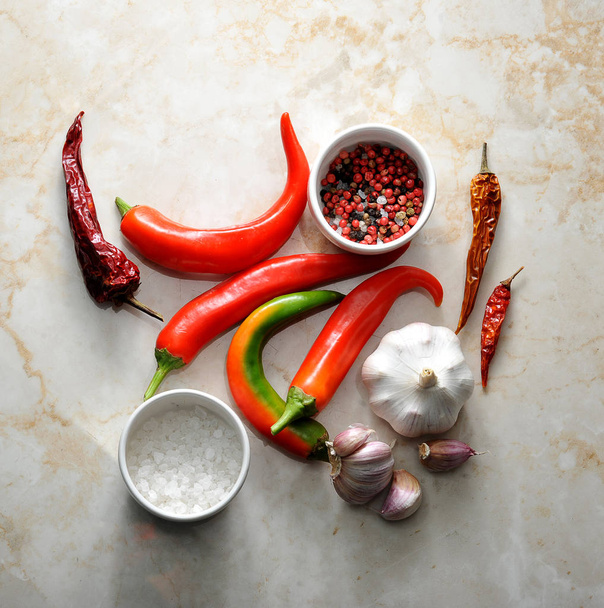 A set of spices. Fresh hot chilli and a few pods of dried pepper. The head of garlic and several separate cloves of garlic. Two cups with salt and pepper. View from above. Light marble background. - Foto, Imagen