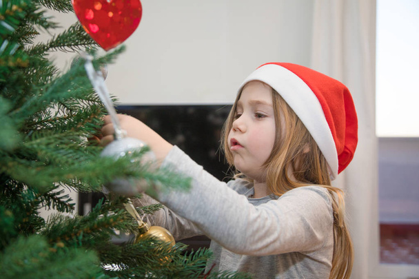 милый маленький ребенок в шляпе Санты, украшающий елку
 - Фото, изображение