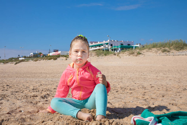 kleines Mädchen, das am Strand sitzt und mit wütendem Ausdruck anschaut - Foto, Bild