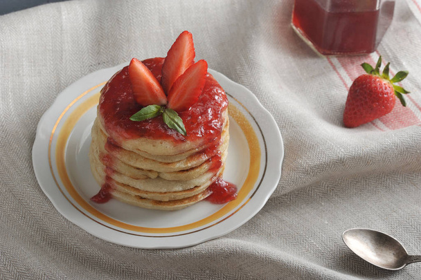 On a plate of pancakes with strawberry jam, decorated with fresh strawberries. The background is draped with a napkin. In the frame, a strawberry, a spoon, a jar of jam. Close-up. Macro photography. - Photo, Image