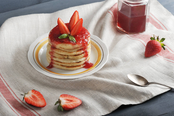 On a plate of pancakes with strawberry jam, decorated with fresh strawberries. The background is draped with a napkin. In the frame, a strawberry, a spoon, a jar of jam. Close-up. Macro photography. - Zdjęcie, obraz