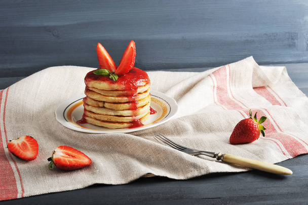 On a plate of pancakes, poured with strawberry jam and decorated with fresh strawberries. In the frame is a fork and a fresh strawberry. Dark background draped with a napkin. Close-up. - Foto, Bild