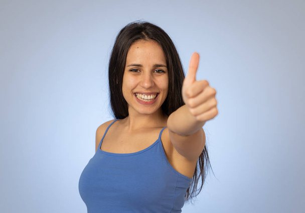 Portrait of beautiful young latin woman showing thumbs up sign feeling cheerful, happy and satisfied. Young happy student woman making thumb up in joyful approval gesture isolated on blue background. - Foto, Bild