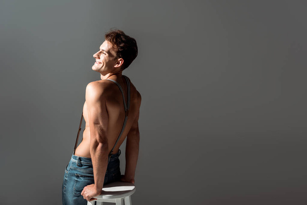 happy shirtless man in suspenders near white chair isolated on grey  - Foto, Imagen