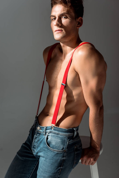 shirtless man in red suspenders looking at camera and standing near white chair isolated on grey   - Fotoğraf, Görsel