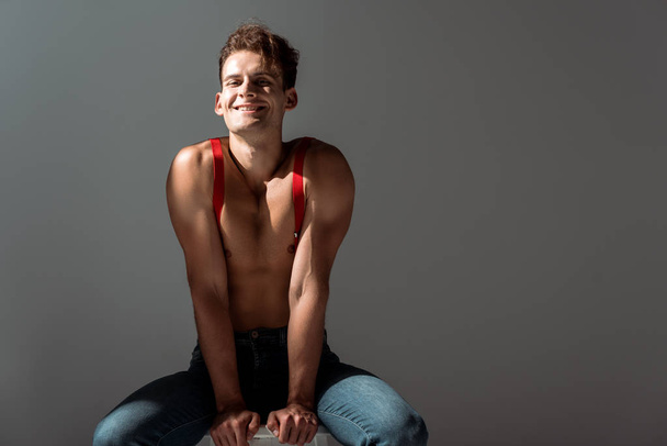 cheerful shirtless man in red suspenders sitting on chair isolated on grey   - Foto, Imagem