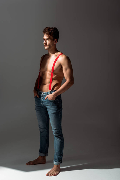 без сорочки і красивий чоловік торкається червоних підтяжок, стоячи на сірому
   - Фото, зображення