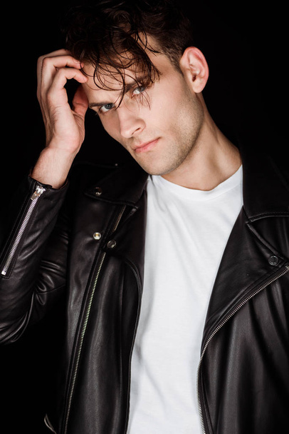 stylish young man in leather jacket touching hair isolated on black  - Foto, Imagem