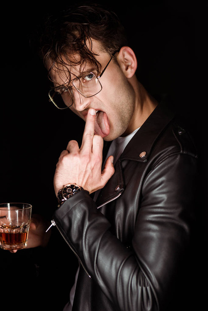 brutal man showing middle finger while sticking out tongue and holding glass of whiskey isolated on black  - Photo, Image