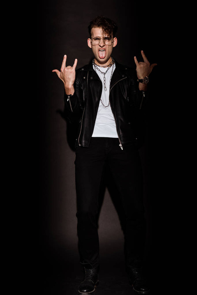 trendy man in glasses showing rock sign while sticking out tongue on black  - Foto, Imagen