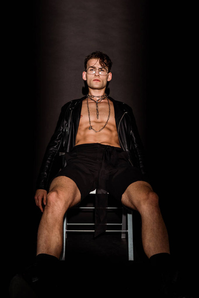 brutal young man in glasses and leather jacket sitting on black  - Φωτογραφία, εικόνα