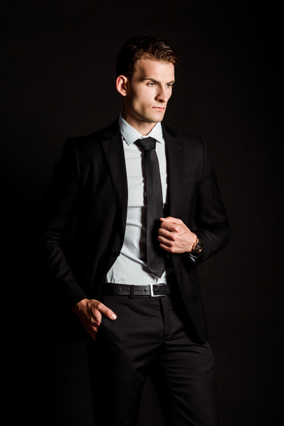 handsome businessman in formal wear standing with hand in pocket isolated on black  - 写真・画像
