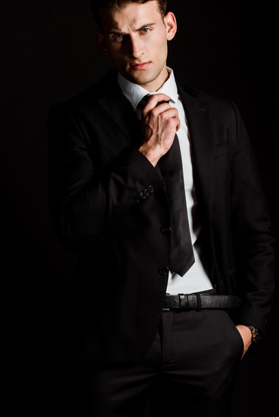 handsome businessman touching tie and standing with hand in pocket isolated on black  - 写真・画像