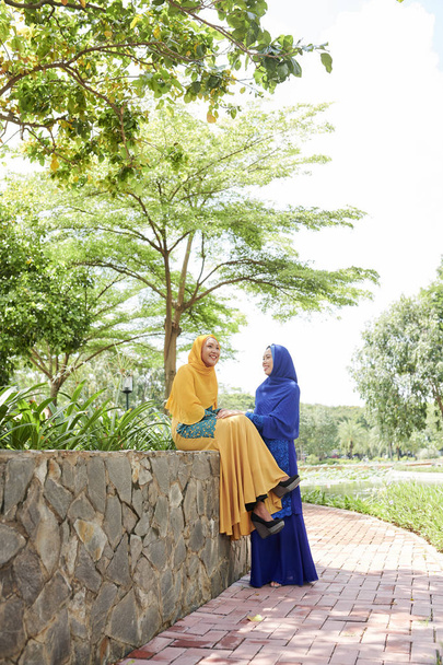 Happy muslim Asian female friends talking and joking when spending summer day together in city park - Photo, Image