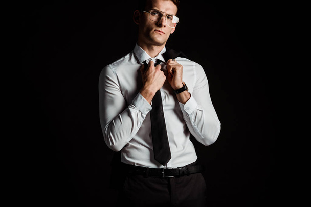 handsome man in glasses touching tie isolated on black  - Фото, зображення