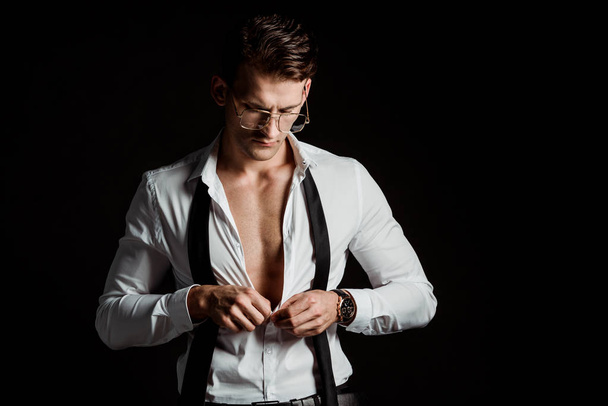 handsome businessman in glasses and formal wear undressing isolated on black  - Fotografie, Obrázek