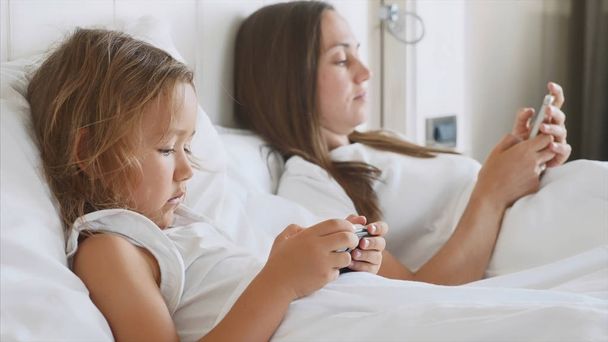 Young mother with little cute daughter are using gadgets lying on a white bed. - Foto, Imagen