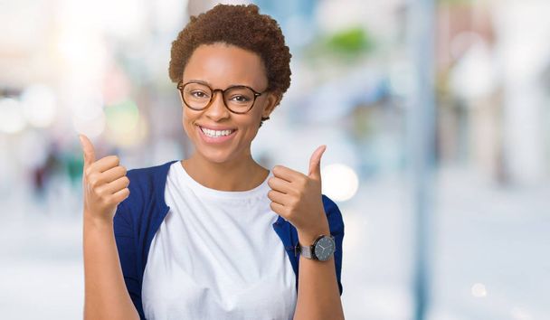 Young beautiful african american woman wearing glasses over isolated background success sign doing positive gesture with hand, thumbs up smiling and happy. Looking at the camera with cheerful expression, winner gesture. - Photo, Image